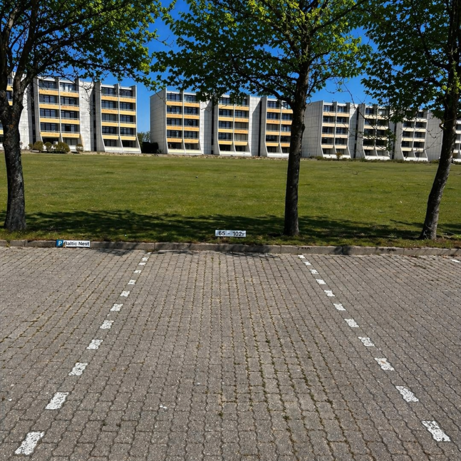 Gepflasterte Parkplätze bei der Meerzeitliebe mit Bäumen, Grünstreifen und Appartementhäusern im Hintergrund.