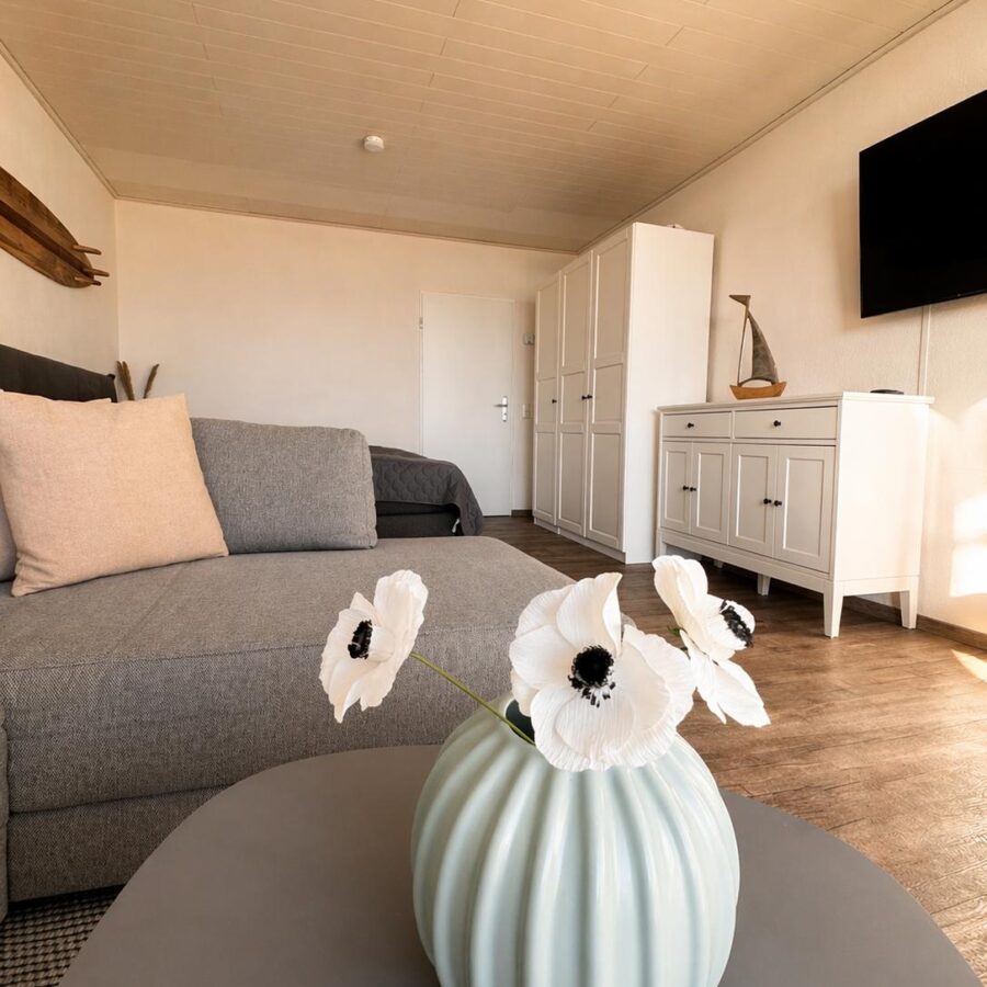 Graues Sofa, weißes Sideboard und Fernseher in der Meerzeitliebe, davor dekorative weiße Vasen.