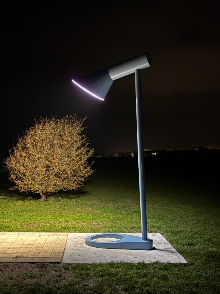 Die blaue AJ-Leuchte bei Nacht auf der dunklen Wiese. Am unteren Rand des schräg gestellten Schirms liegt ein heller Lichtstreifen, davor ein angestrahlter Strauch.
