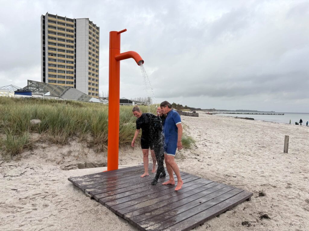 Drei Menschen in Badekleidung stehen barfuß auf dem Holzdeck unter der orangefarbenen Stranddusche im Wasserstrahl, dahinter Dünengras und die Ostsee.