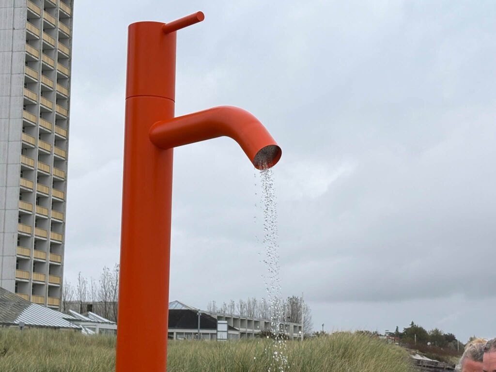 Nahaufnahme der orangefarbenen Stranddusche in Form einer Armatur: der gebogene Auslauf, aus dem Wasser fällt, und der aufrechte Griff daneben.