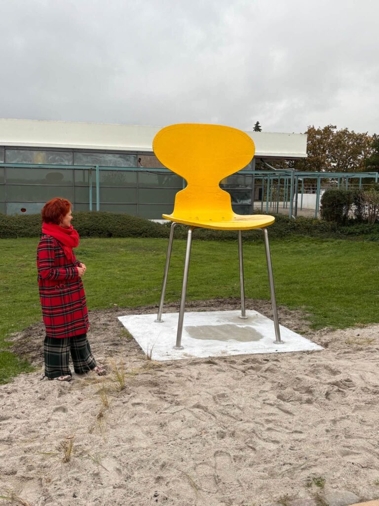 Der gelbe XXL-Ameisenstuhl auf einem Betonsockel im Sand, daneben steht Gabriele Staarmann im roten Mantel. Die Sitzfläche reicht ihr etwa bis zur Schulter.