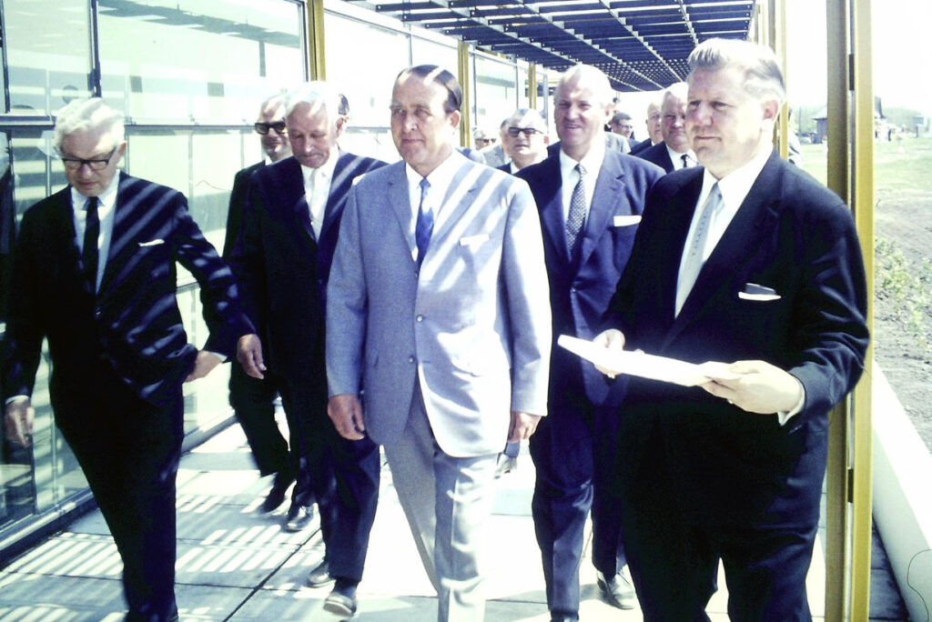 Eine Gruppe von Männern in Anzügen geht durch den verglasten Wandelgang des neuen Gebäudes in Burgtiefe. Links außen Arne Jacobsen.